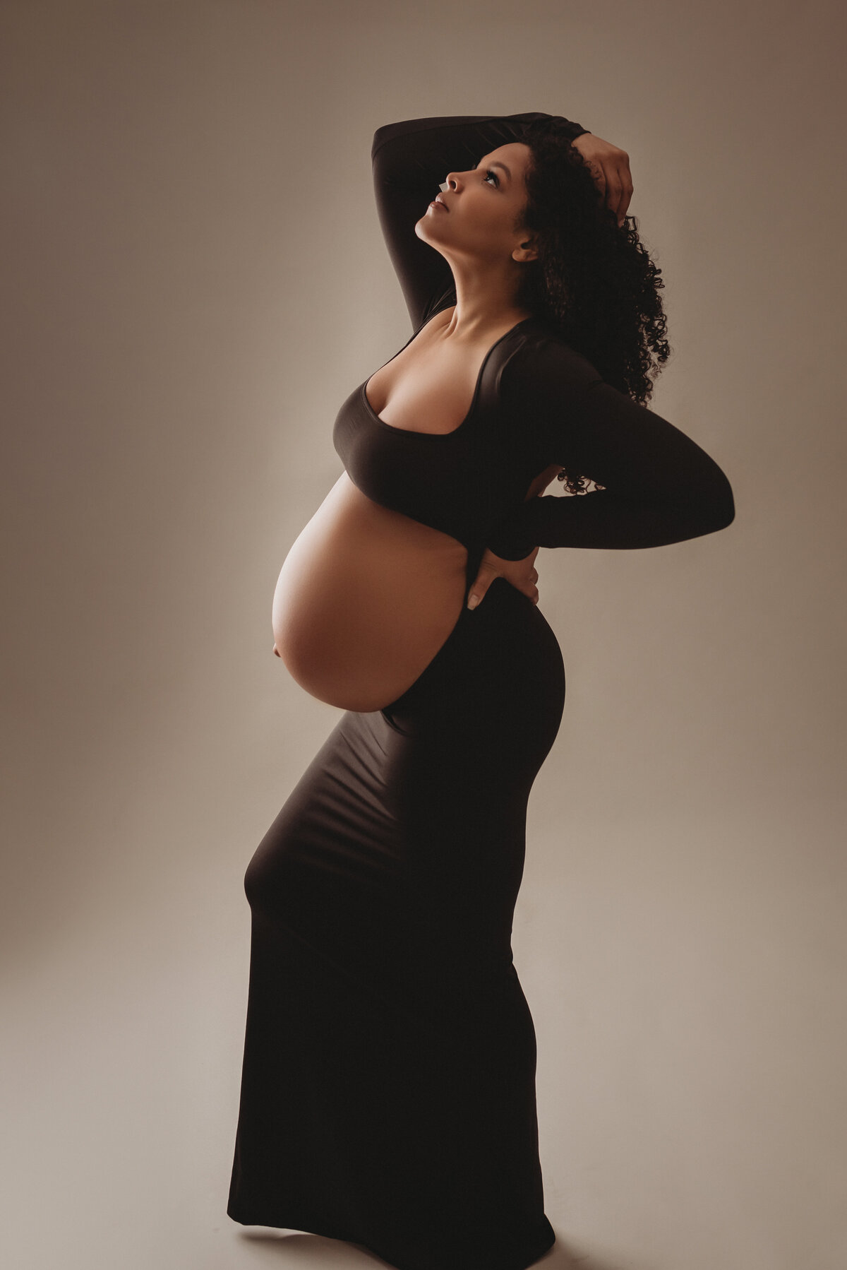 High end maternity photo shoot in Atlanta with Marietta, GA maternity photographer showing 36 week pregnant woman dressed in a black maternity gown showing her baby bump posing on a light gray backdrop.