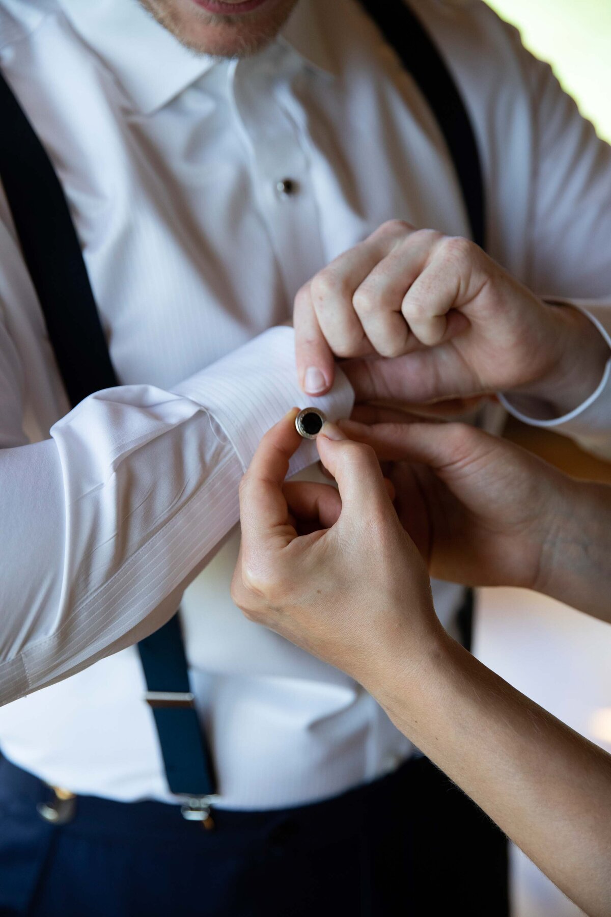 Detailed image of a groom receiving help with his cufflinks. Groomsman in suspenders getting ready at Zigbone Farm Retreat, Maryland