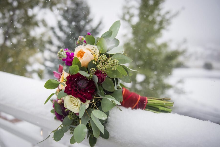 A. bouquet of various red, pink and cream flowers sits on a snowy porch ledge, captured by Denver wedding photographer, Two One Photography.