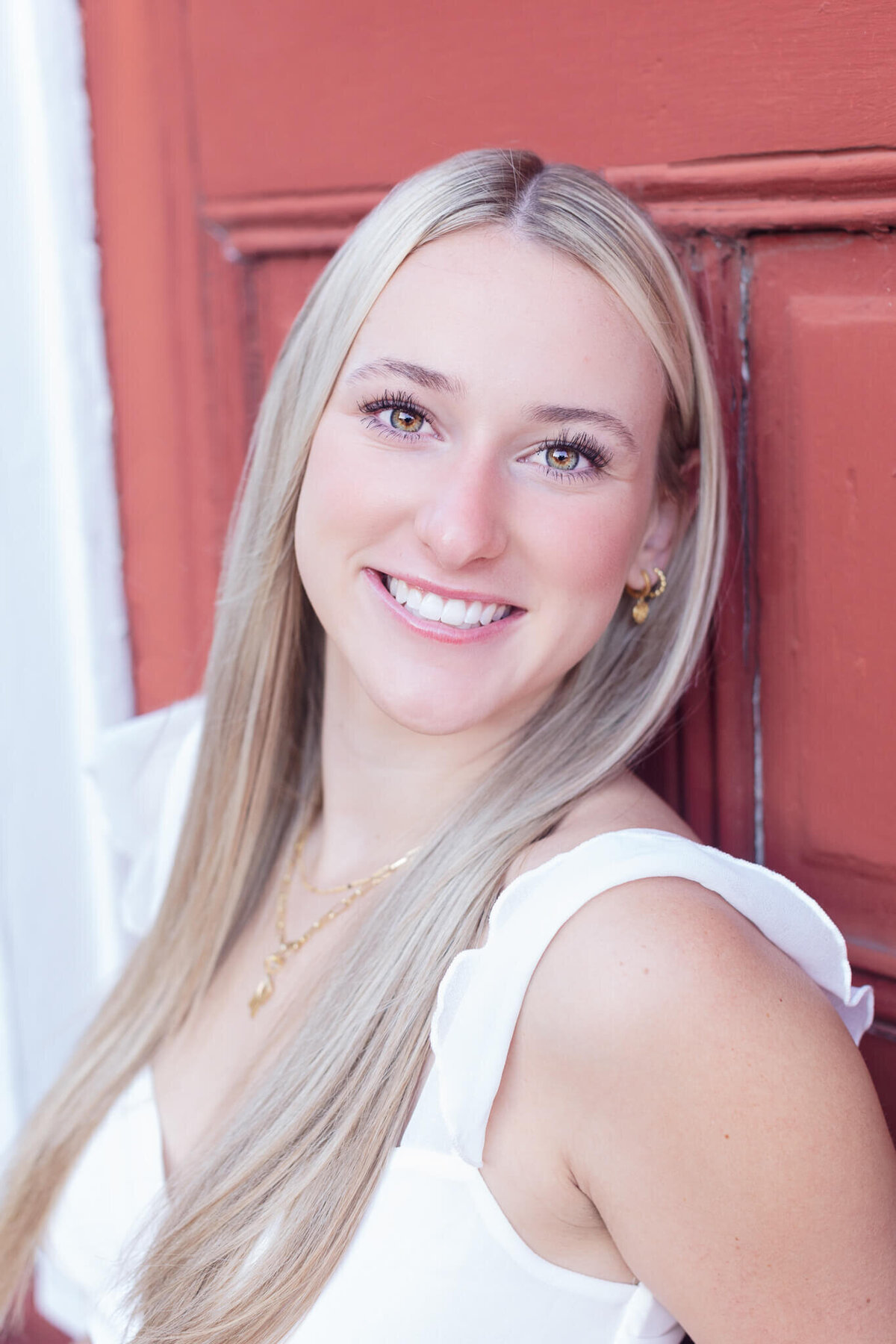 A person with long blonde hair, smiling, stands in front of a red door. They are wearing a white top and gold earrings.