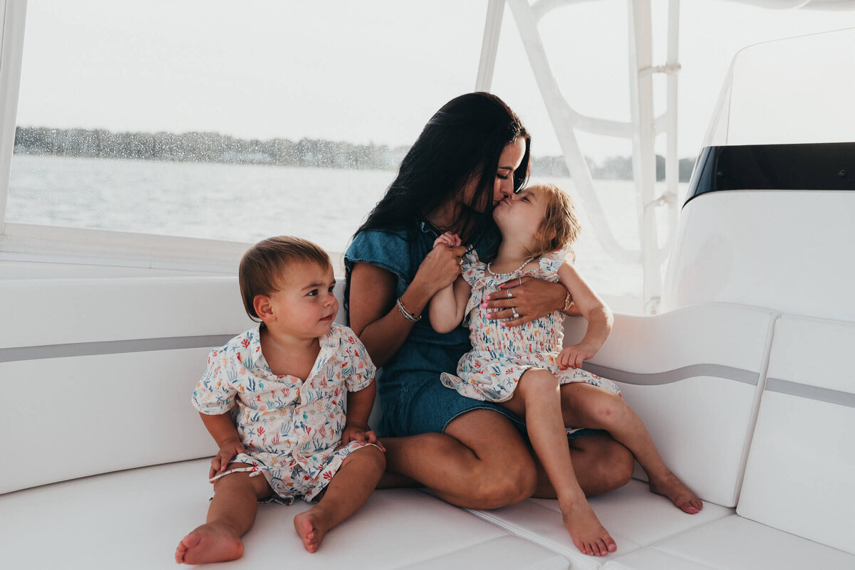 A mother sits with her two young children on their family boat, her daughter is sitting on her lap and giving her a kiss