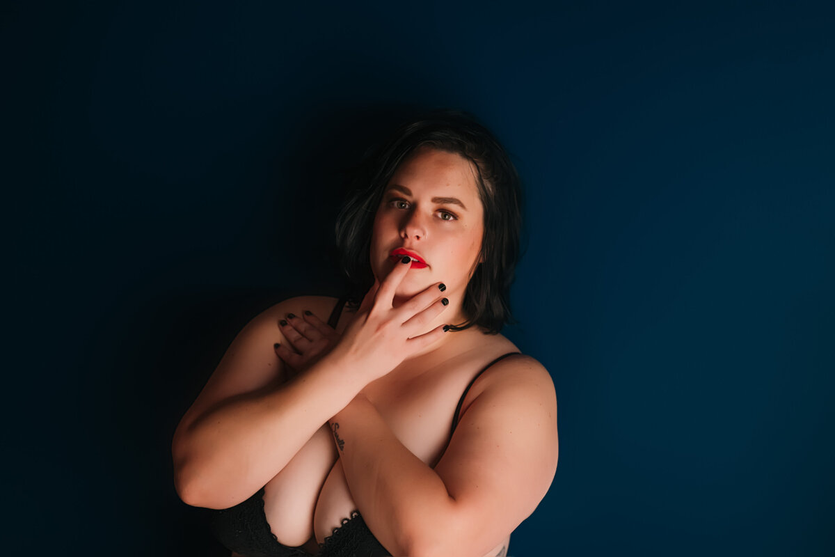 lady with black hair and red lipstick touching her lips against a blue wall for her boudoir session in Nashville