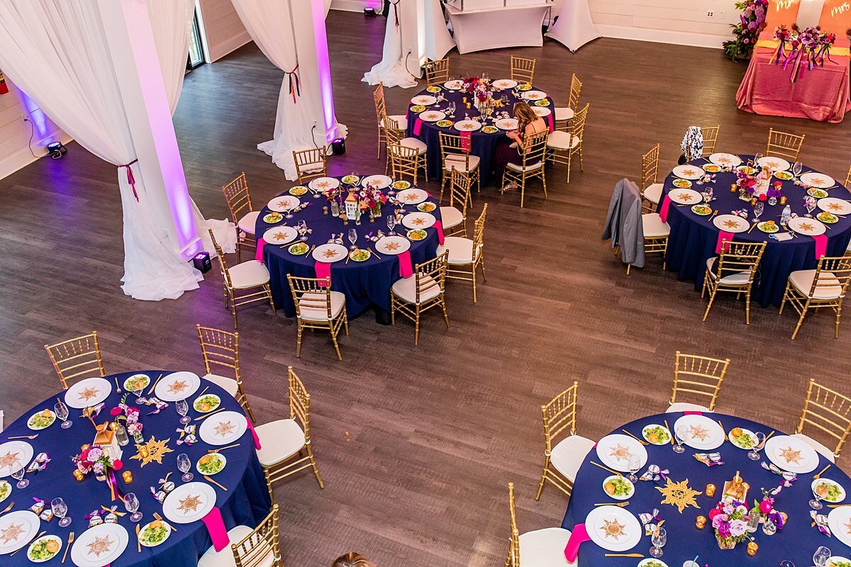 Ballroom at Sugarfoot set for a Tangled themed wedding with royal blue table cloths, hot pink napkins, gold chivari chairs, white plates with gold suns and gold lanterns with hot pink and purple flowers.