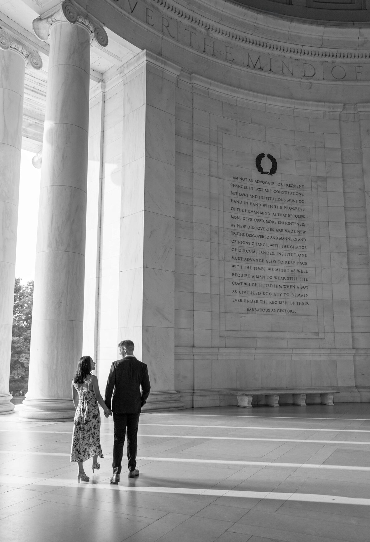 A couple walks hand in hand inside a grand, columned monument. The tall, engraved walls feature inscriptions, and light streams in from an open side, casting shadows on the floor. The scene conveys a sense of history and intimacy.