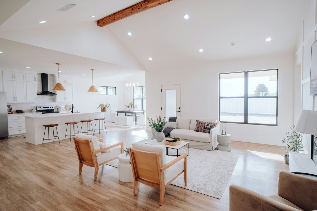A bright, open-concept living space and kitchen in a modern home. The foreground features two armchairs with light wooden frames and white cushions, arranged on a light-colored rug. Behind them, a white sofa faces a large window. Further back, a contemporary kitchen with white cabinetry, a large island with three bar stools, and pendant lighting is visible. Light wooden floors extend throughout the entire space, giving it a clean and airy feel.