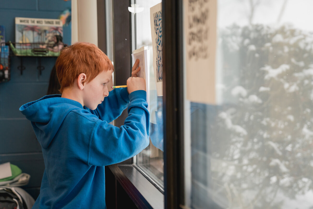 Red-headed middle school boy, tracing a paper against the window of his classroom