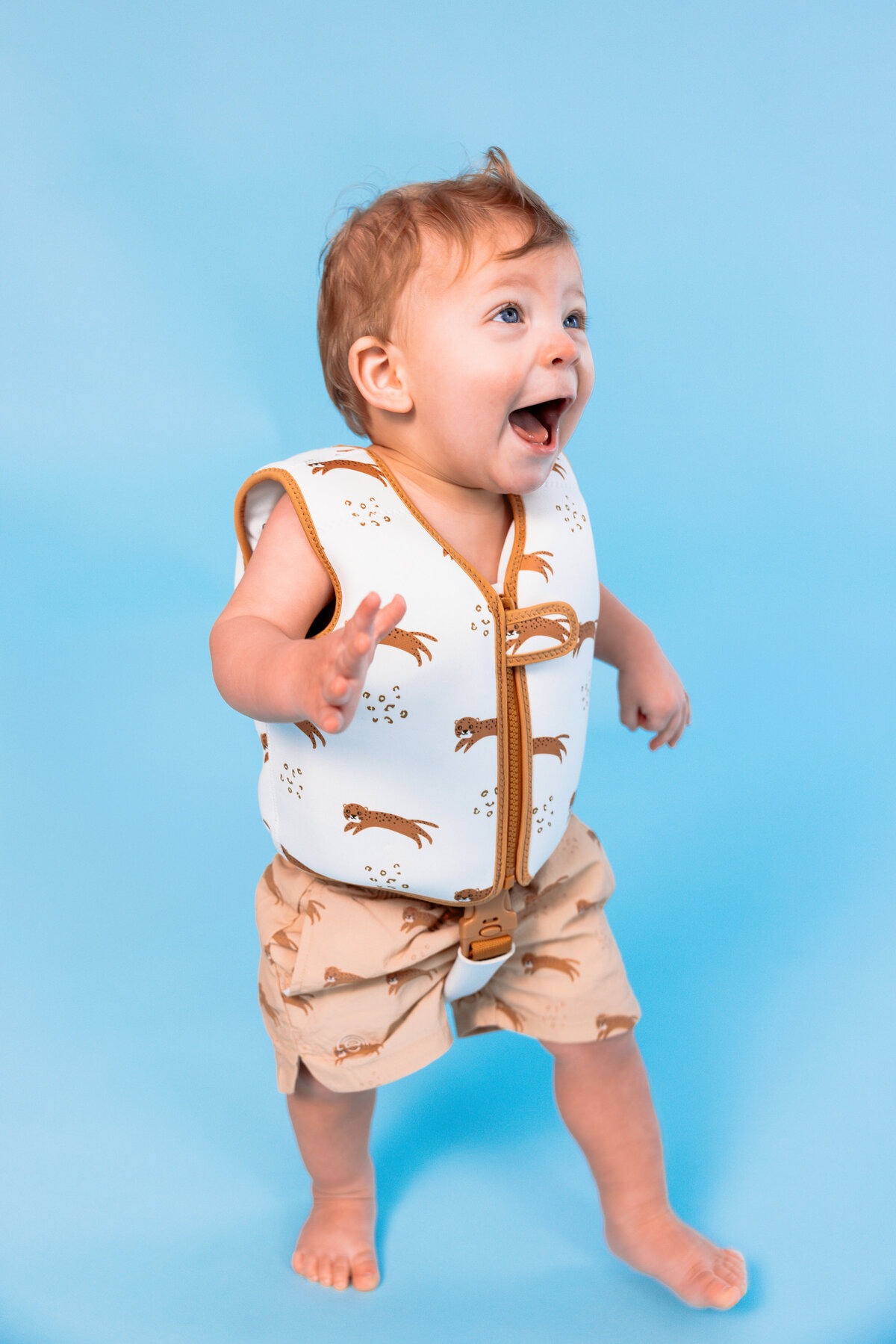 Happy toddler wearing Swim Essentials beige float vest and matching swim shorts with panther print, standing against a light blue background.