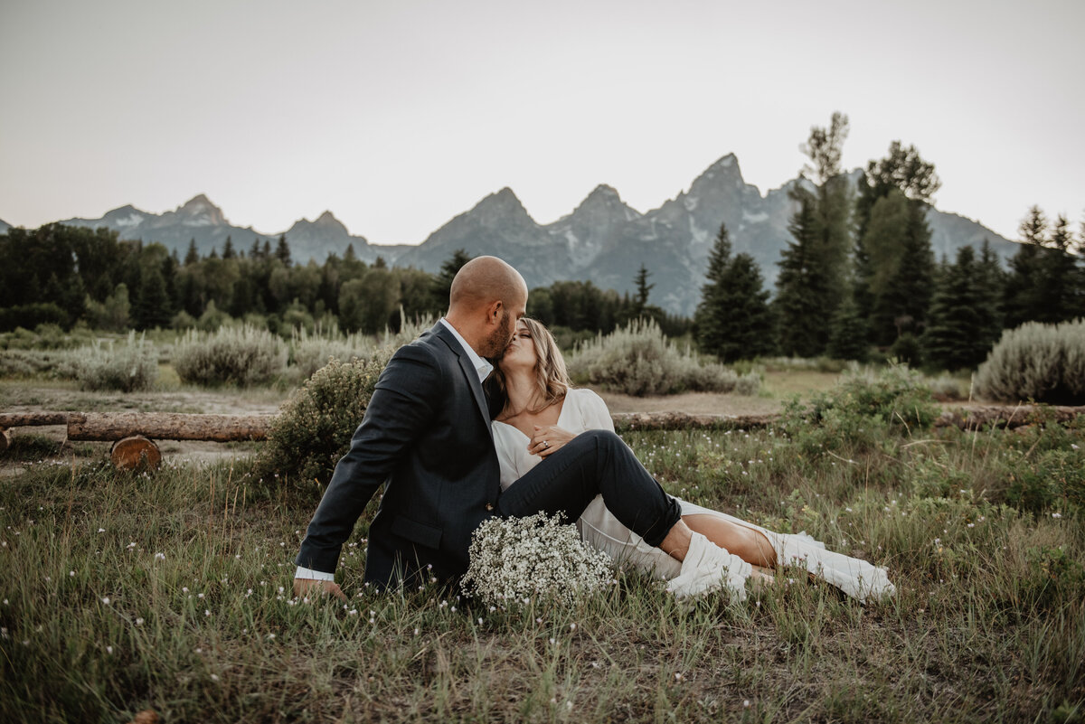 grand Teton elopement with bride and groom sitting in a field in front of the mountain range kissing photographed by jackson wyoming photographer