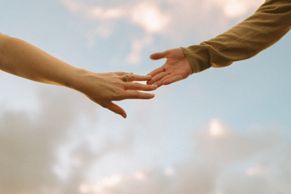 a photo of a couples hands barely touching with the cotton candy colored sky in the background.