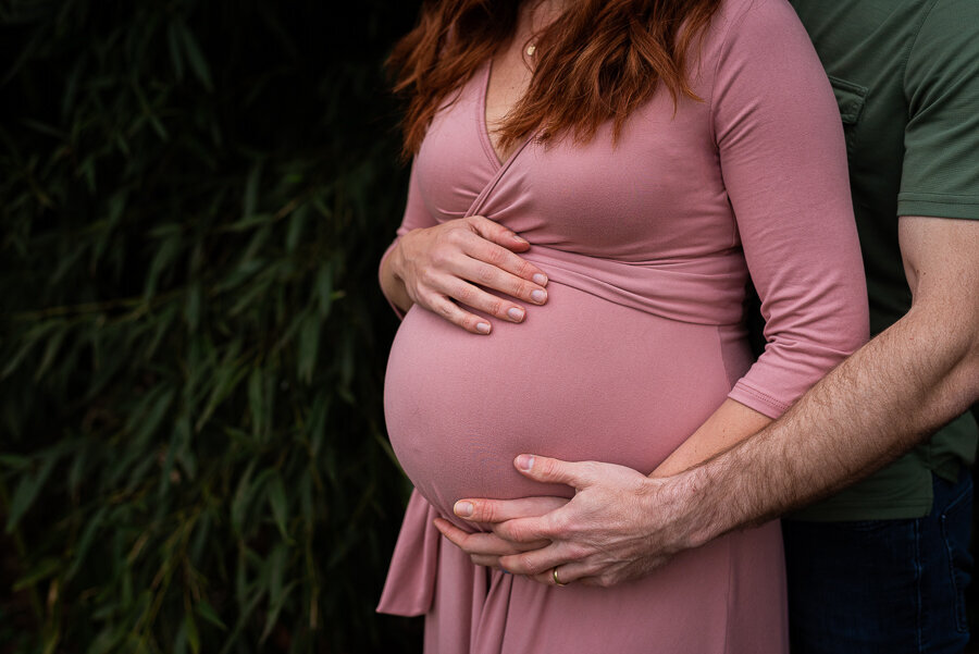 A woman in a pink dress and her husband hold her pregnant belly.