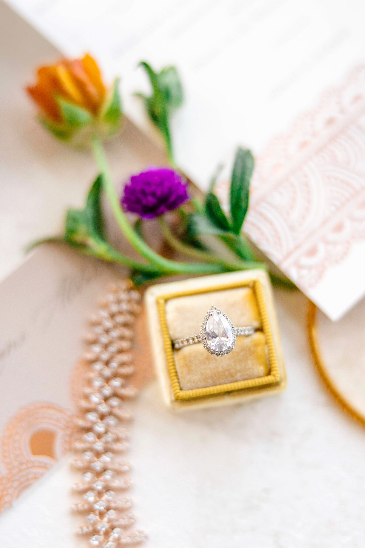 A close-up of a pear-shaped diamond ring in a yellow velvet box, surrounded by delicate flowers and decorative stationery on a white surface. A sparkling bracelet is also visible on the left side.