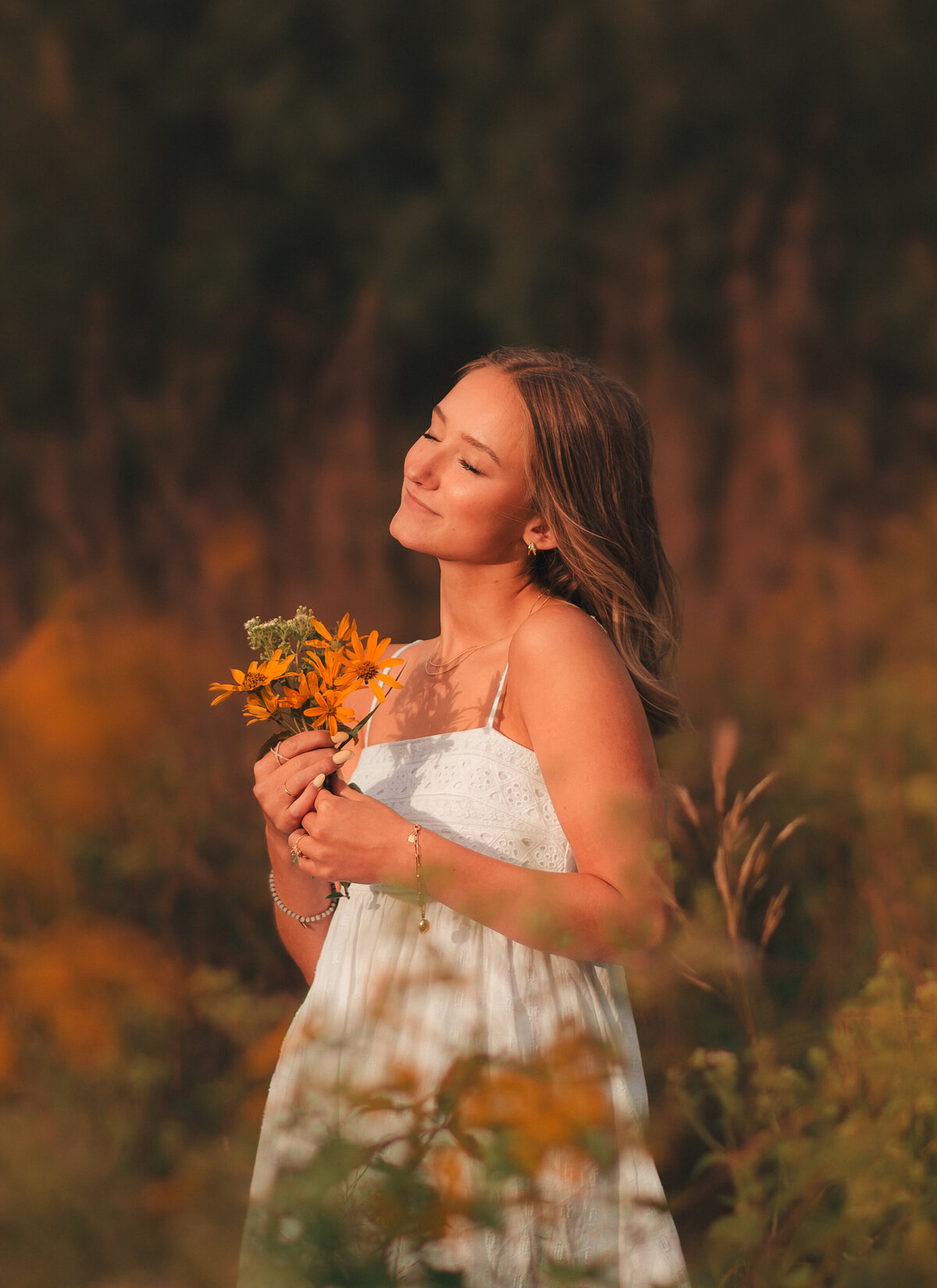 High school senior in flowy white dress walking through grassy field at sunset in Minnesota.
