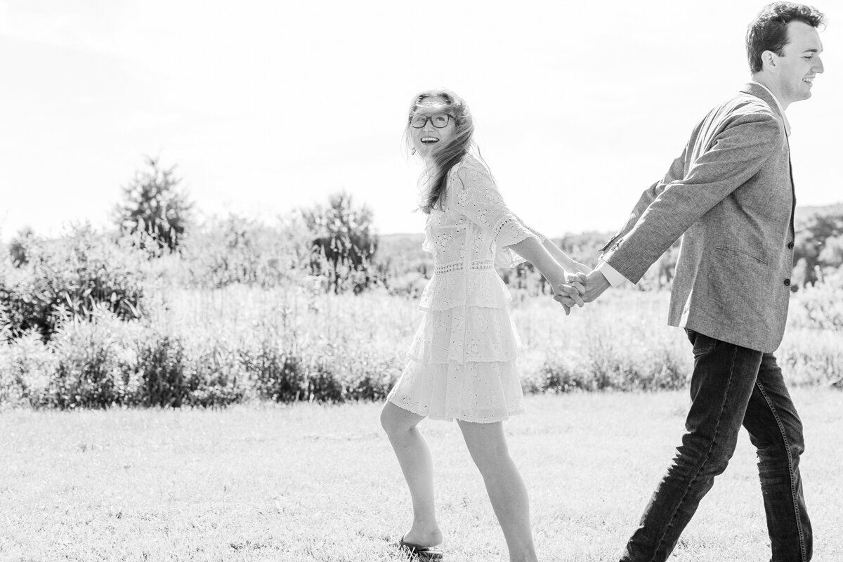 Candid black and white photograph of engaged couple spinning in a field in Cooperstown, NY