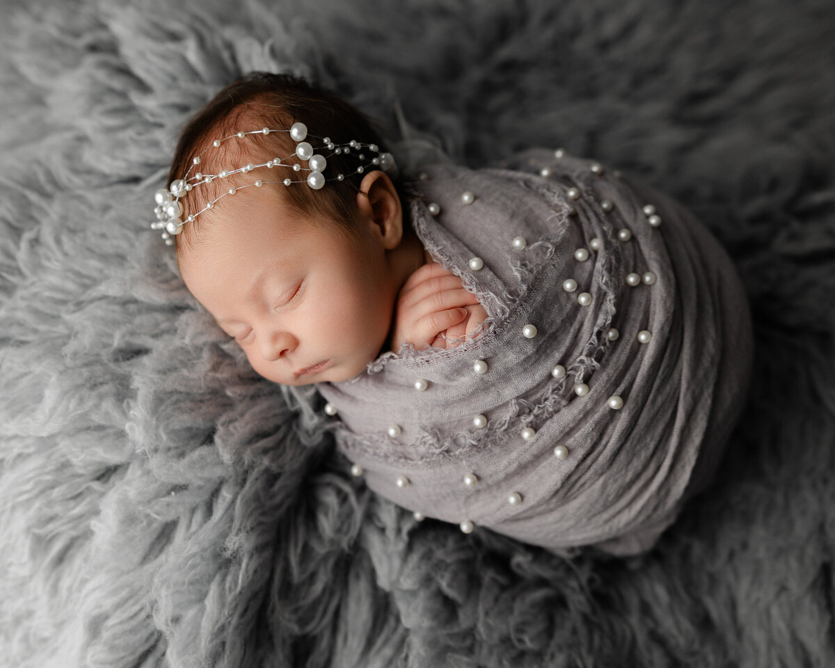 Baby girl peacefully sleeping in a pearl-accented swaddle with a matching pearl headpiece, posed on a cloud-like gray fur layer. Photographed by Inland Empire newborn photographer Kaitlyn Dawn.