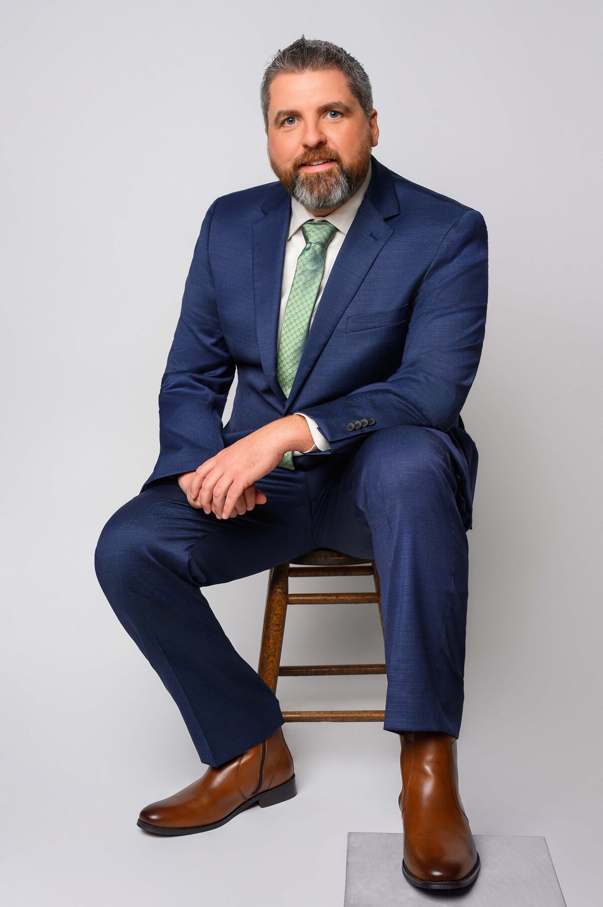 Man in a blue suit and green tie, sitting on a wooden stool