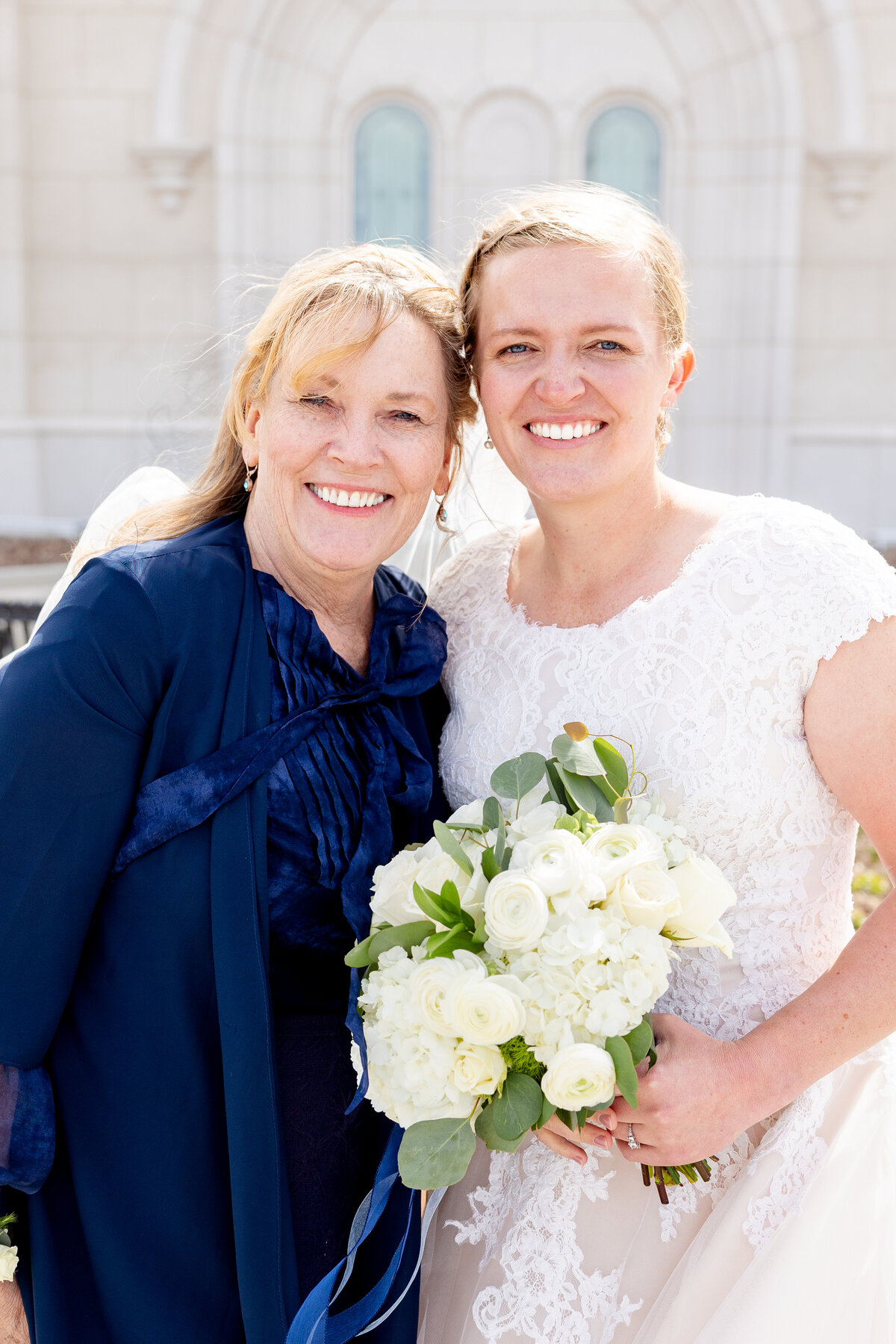 The bride stands close with her mother, both smiling warmly and holding a bouquet of white flowers outside the temple.