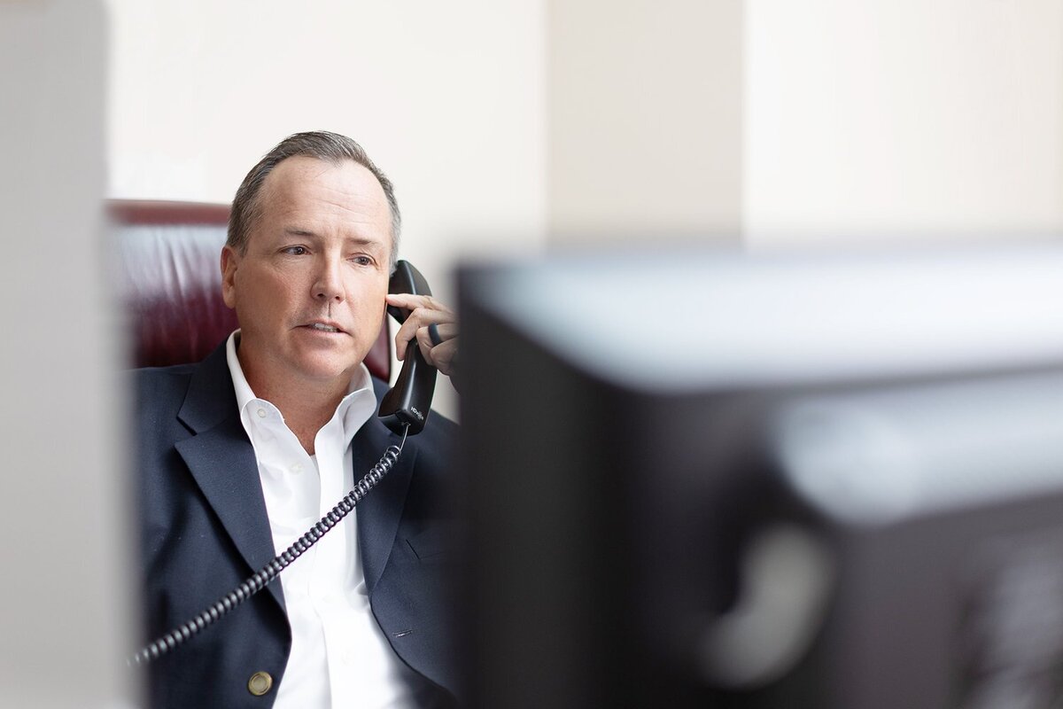 older man in a suit talking on the phone with the corner of a computer out of focus in the foreground