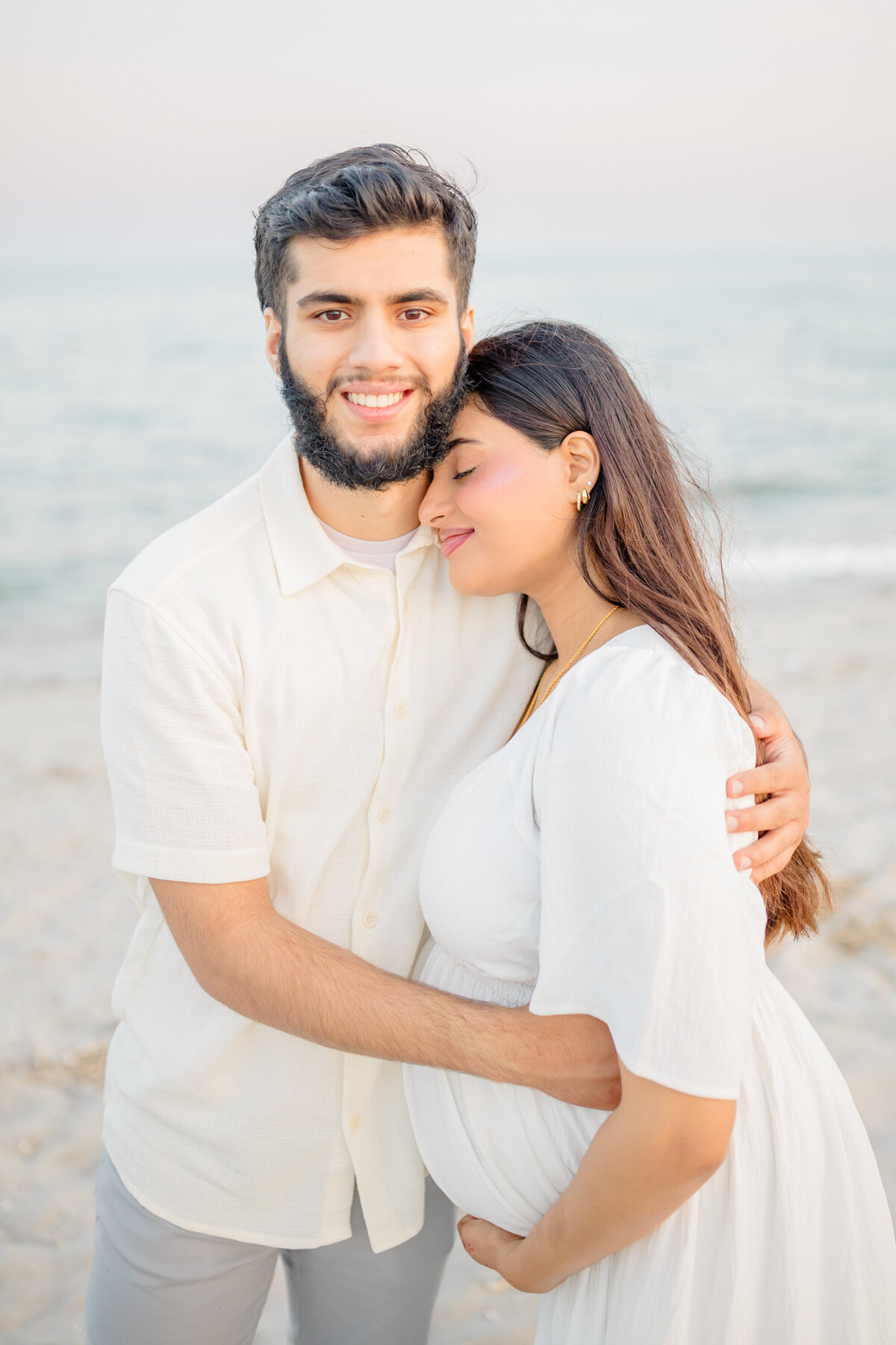 Maternity photo of a couple with the mom-to-be standing on a beach with a flowing white gown and the golden sun setting in the background
