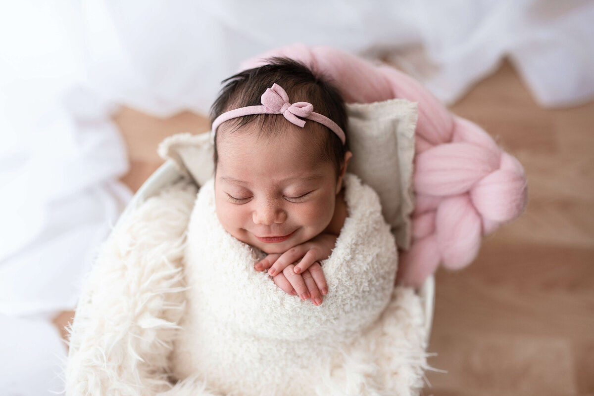 Smiling newborn baby girl wrapped up in bed, with red hair, a headband with a bow and floral swaddle.