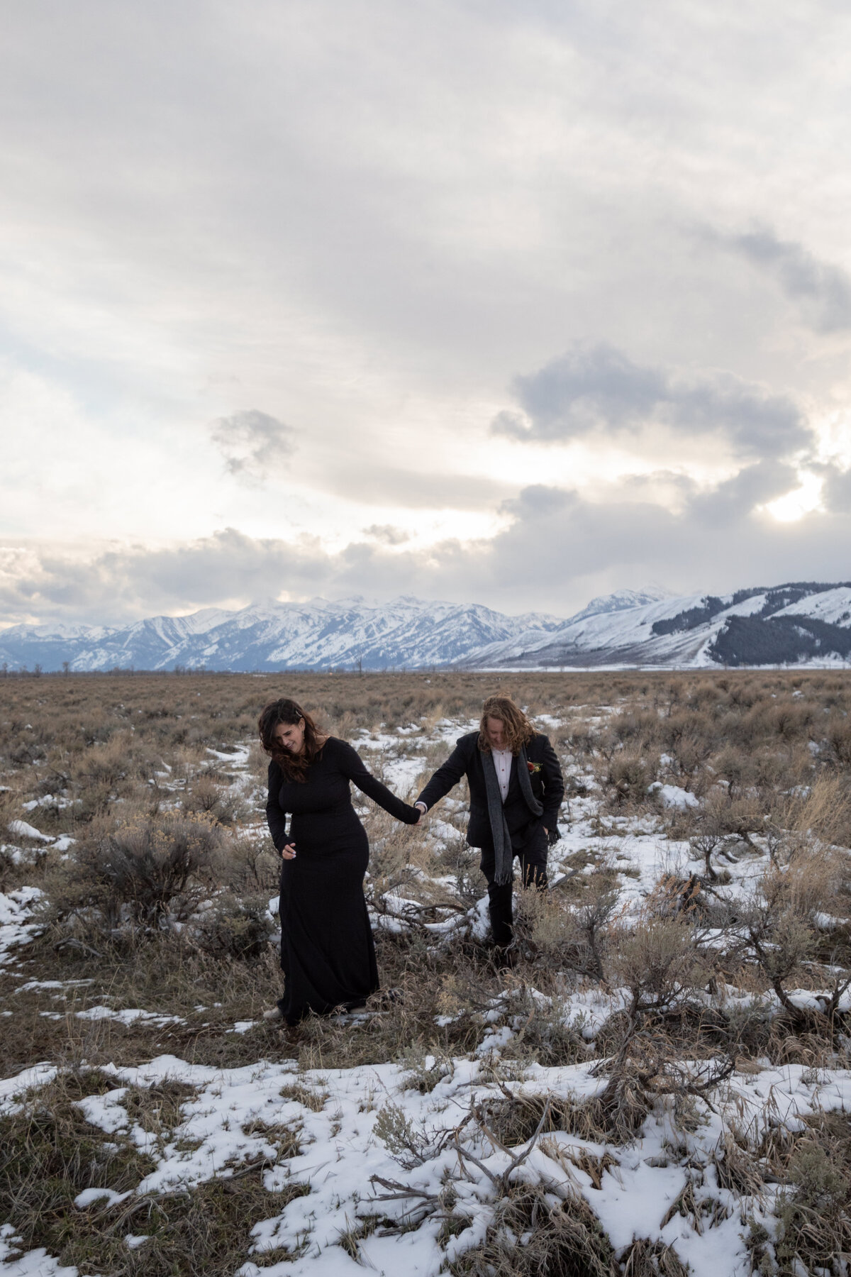 Two brides wearing black walk hand in hand through a snowy meadow during their elopement in Grand Teton.