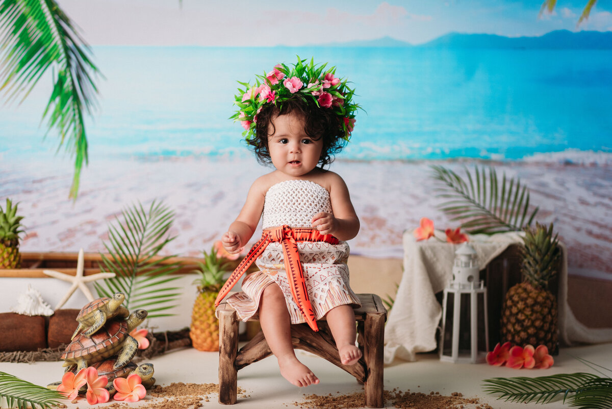 One year baby girl dressed up as Moana sitting in little chair on beach backdrop