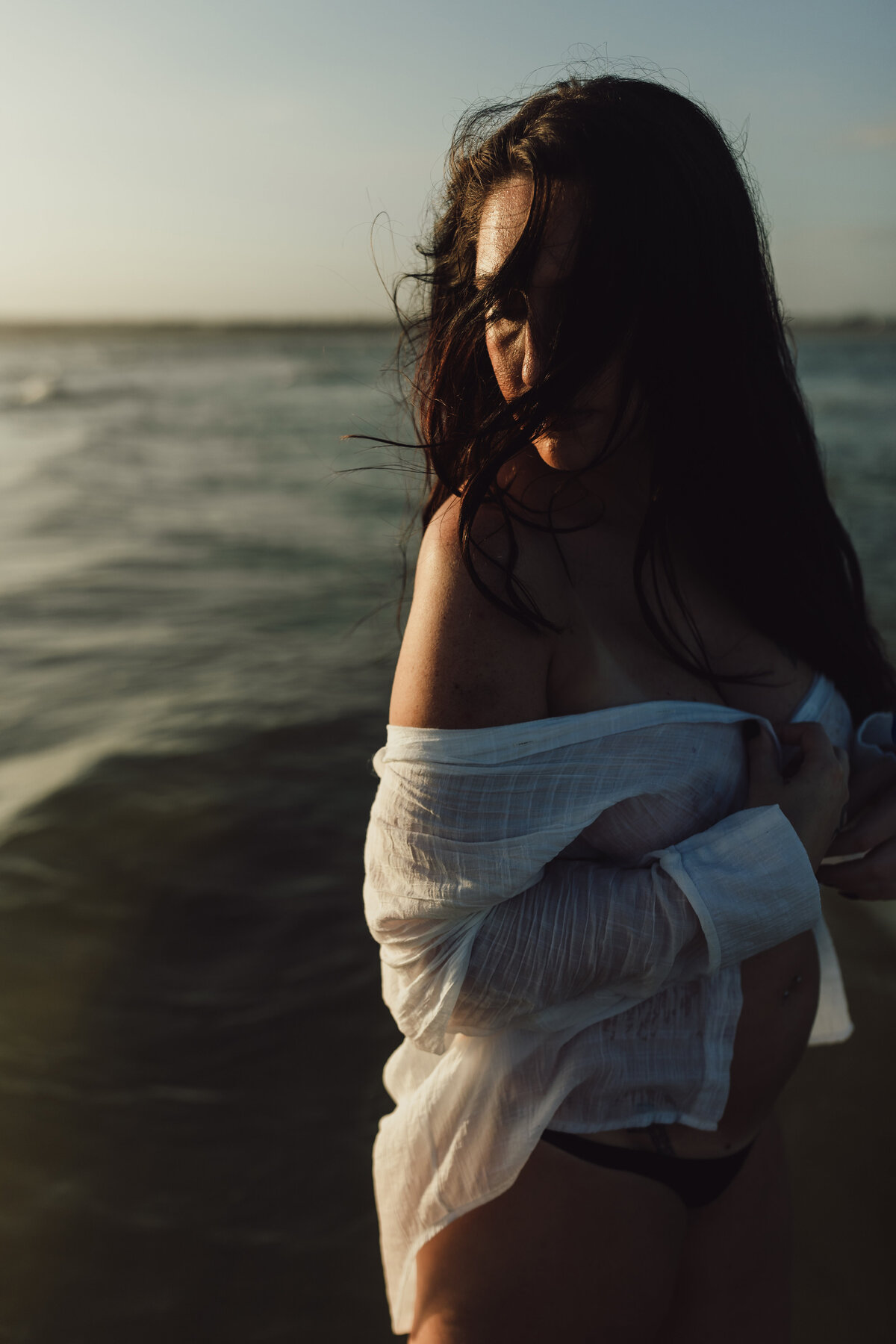 Woman wrapped in a white shirt stands in ocean waves, her hair windswept and face partially hidden during golden hour on the beach.