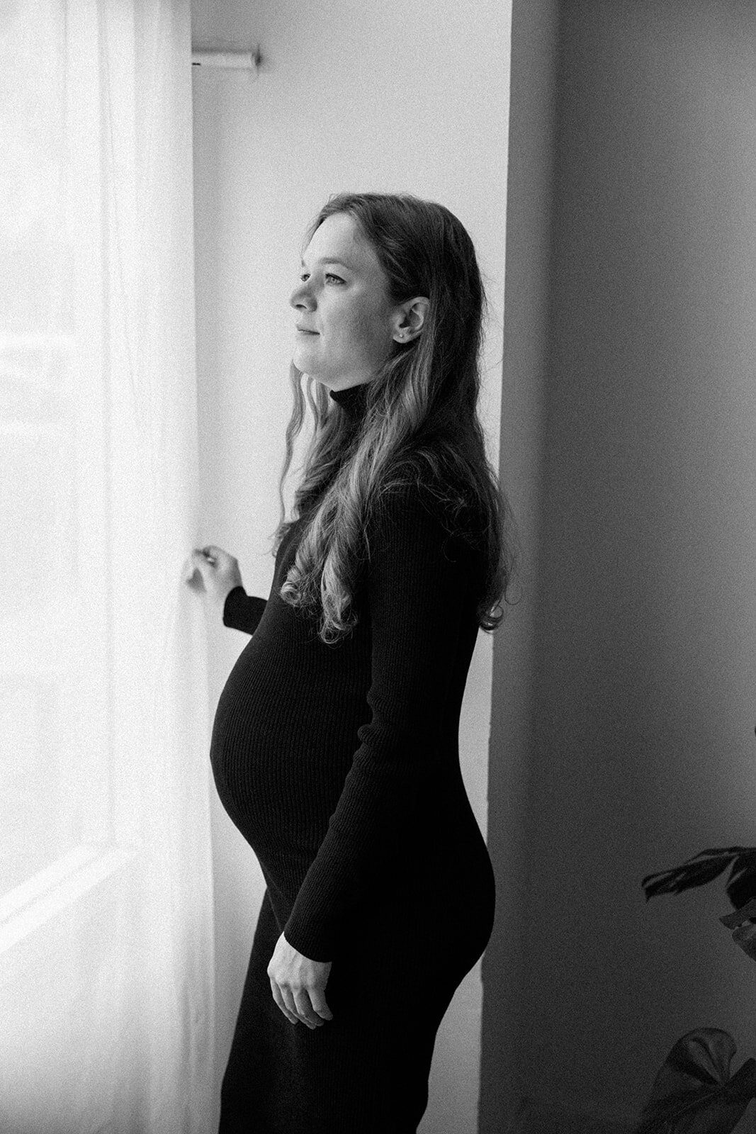 Pregnant woman gazes out the window showing her fully belly. 