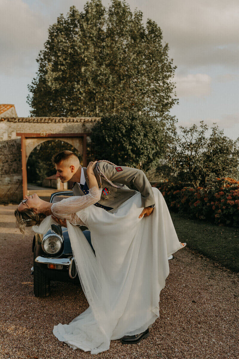 Marié en uniforme basculant en arrière la mariée devant une voiture noire ancienne pour le shooting photo mariage en vendée.