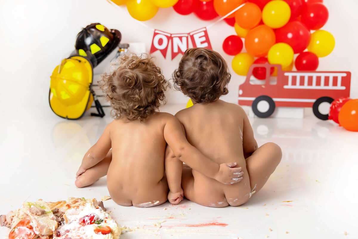 Two curly-haired babies sit on a white floor, surrounded by colorful balloons and a fire truck decoration. A "ONE" banner hangs above, with cake smeared beside them—a perfect capture for Jacksonville cake smash photography.