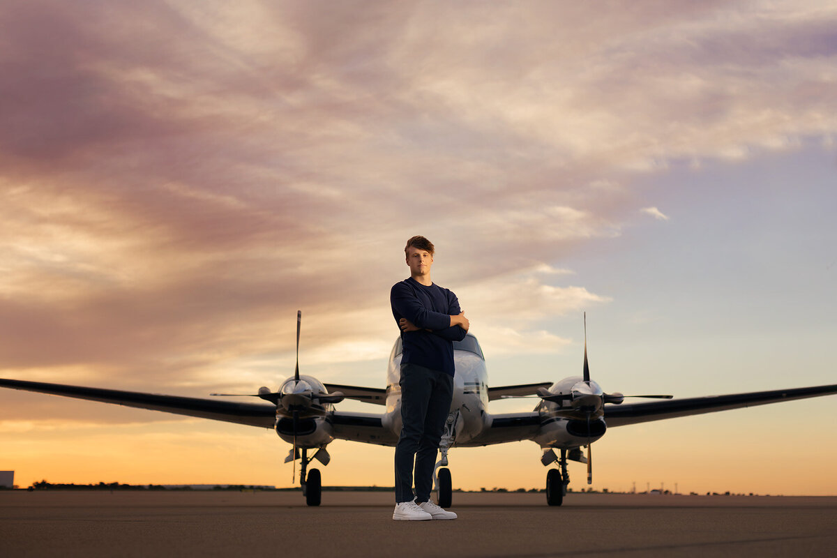 A senior poses confidently in front of a vintage airplane at sunset, showcasing their adventurous spirit. Captured by a Dallas senior photographer.