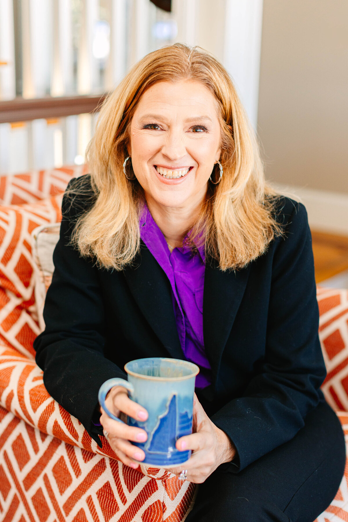 A woman with shoulder-length blonde hair and hoop earrings sits on an orange-patterned chair, smiling warmly while holding a blue ceramic mug. She wears a black blazer over a purple blouse, creating a bold contrast. The cozy indoor setting suggests a relaxed yet professional atmosphere