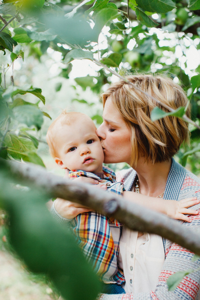 Mother kisses baby on the cheek framed by greenery