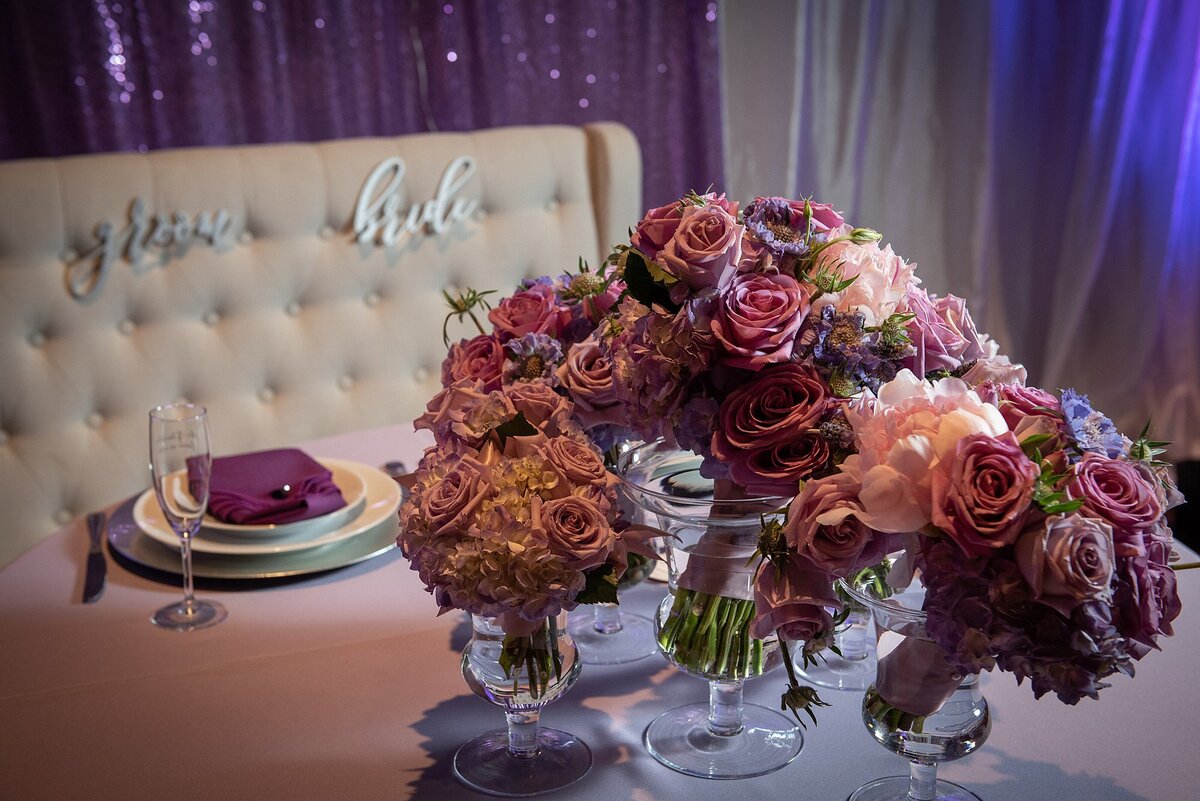 The purple sweetheart table sits in front of a  white  leather love seat  with silver Bride and Groom signs. The bride's bouquet of purple roses, pink roses and light purple roses sits in a glass vase and is surrounded by the bridesmaids light purple rose bouquets.