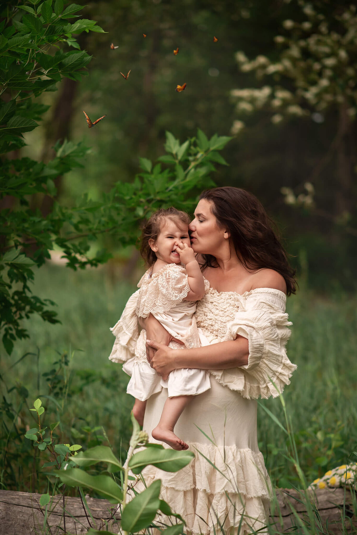 A mother and daughter engage in a loving, silly embrace. Mom kisses her cheek while holding her on her hip. Daughter laughs while butterflies surround them. Captured by Brittany Danielle