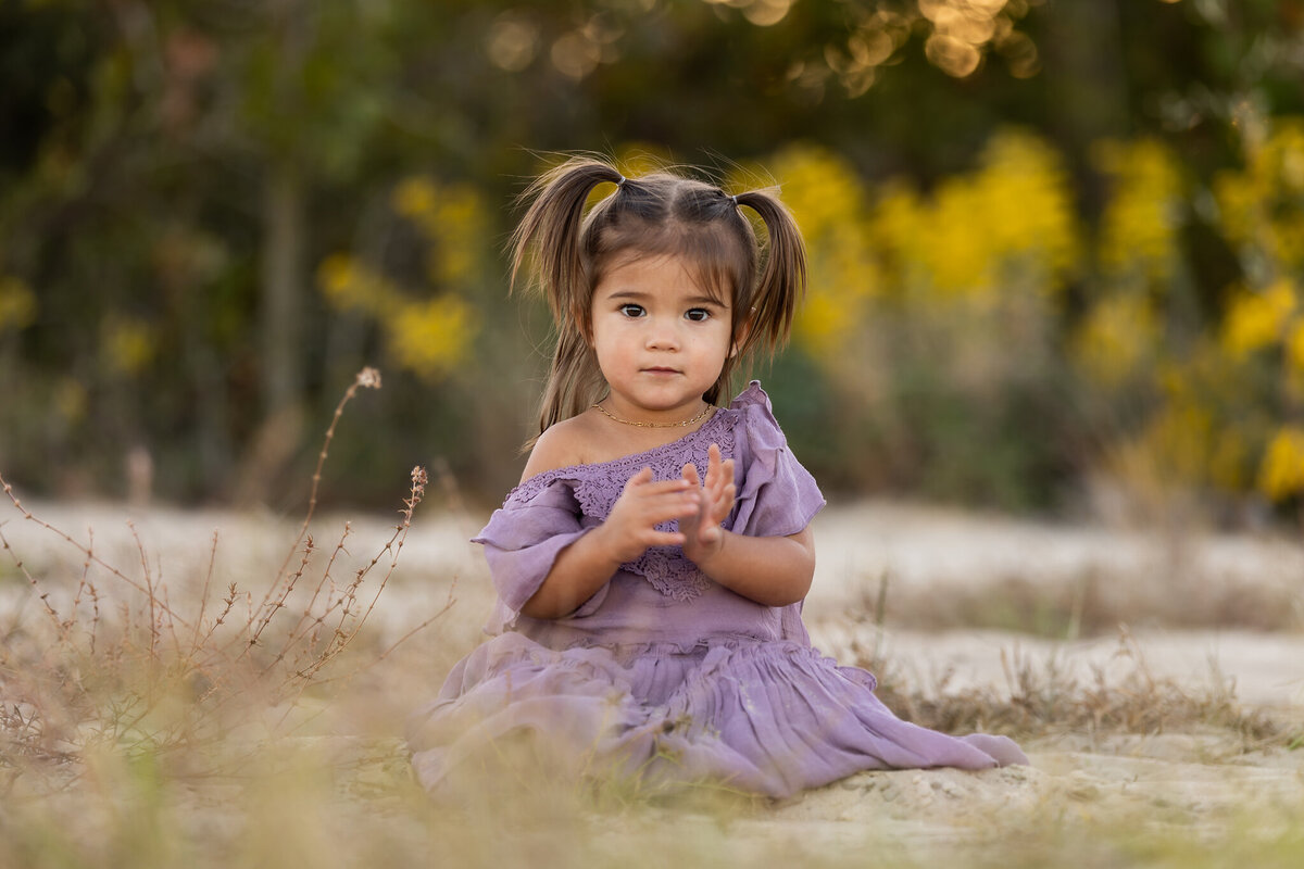 precious little girl with brown hair and brown eyes in a purle dress sitting on ground.