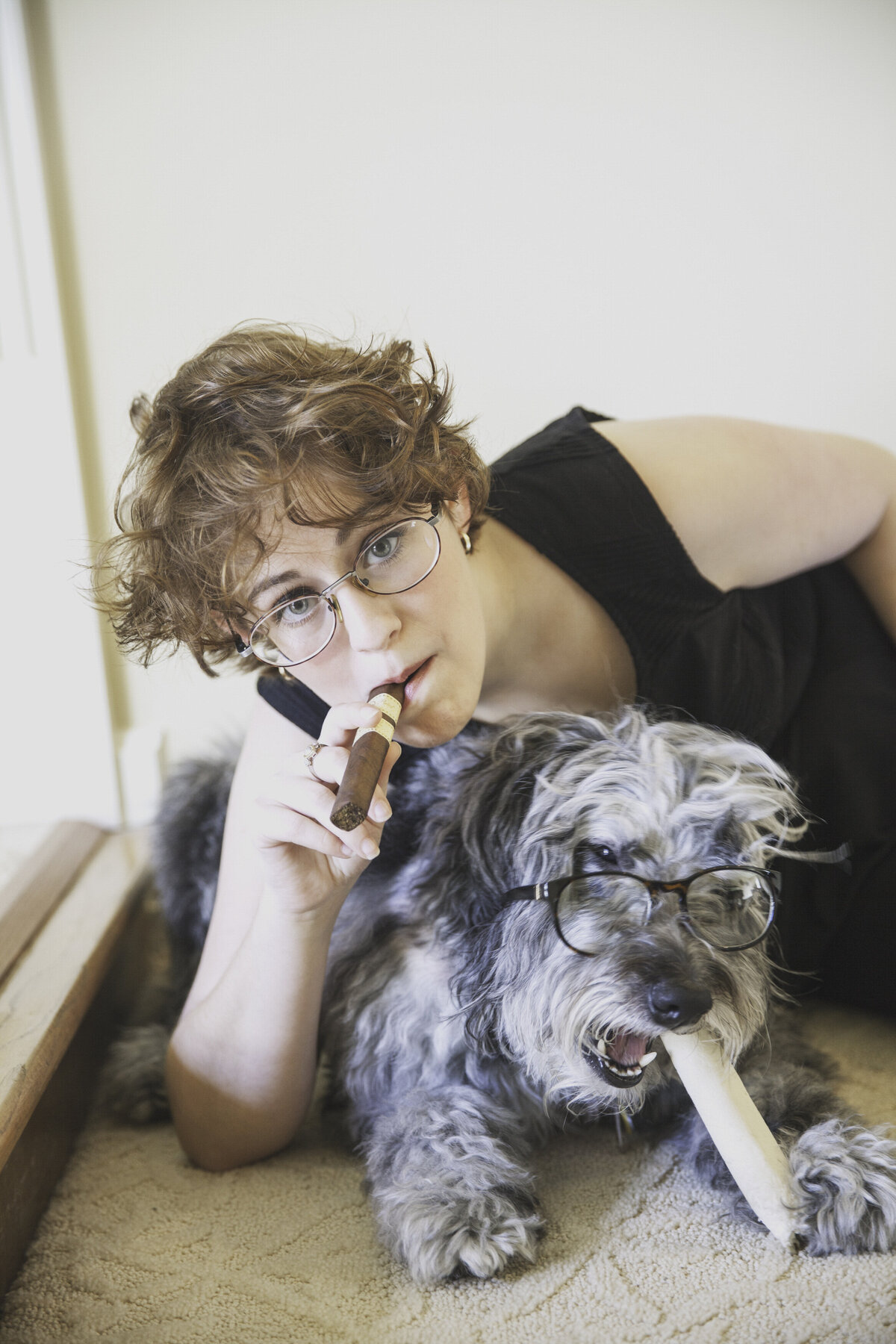 A person holds a cigar while leaning over their dog who wears sunglasses.