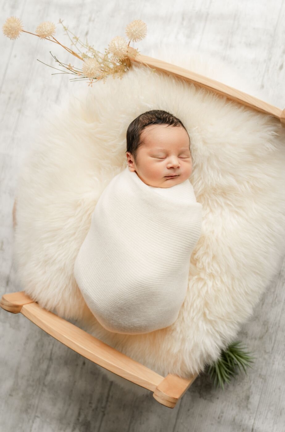 Newborn baby swaddled in white, sleeping on a fluffy cream rug in a small wooden bed decorated with dried flowers.