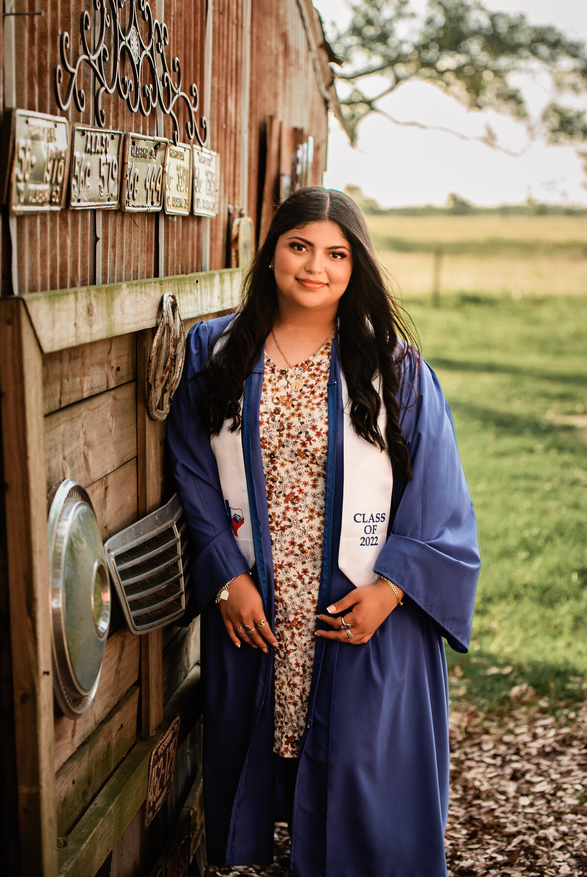 A high school senior wearing her blue gown leans against a blue building.