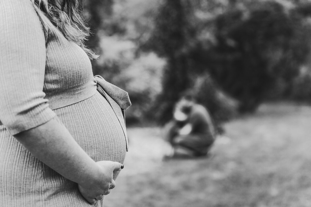 black and white maternity photo. Closeup of baby bump with family in the background
