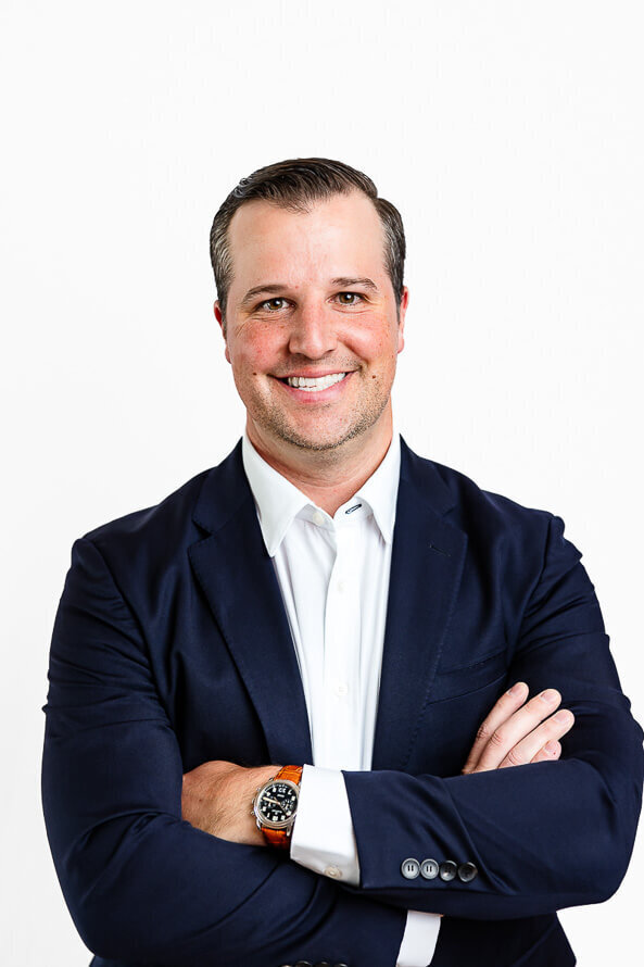 Man in blue coat folds arms and smiles at camera in a white photography studio.