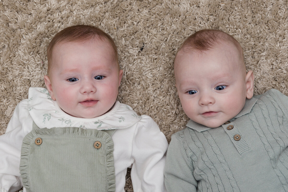 Two infants lying side by side on a beige carpet, one dressed in a white and green outfit, the other in a green outfit with buttons.