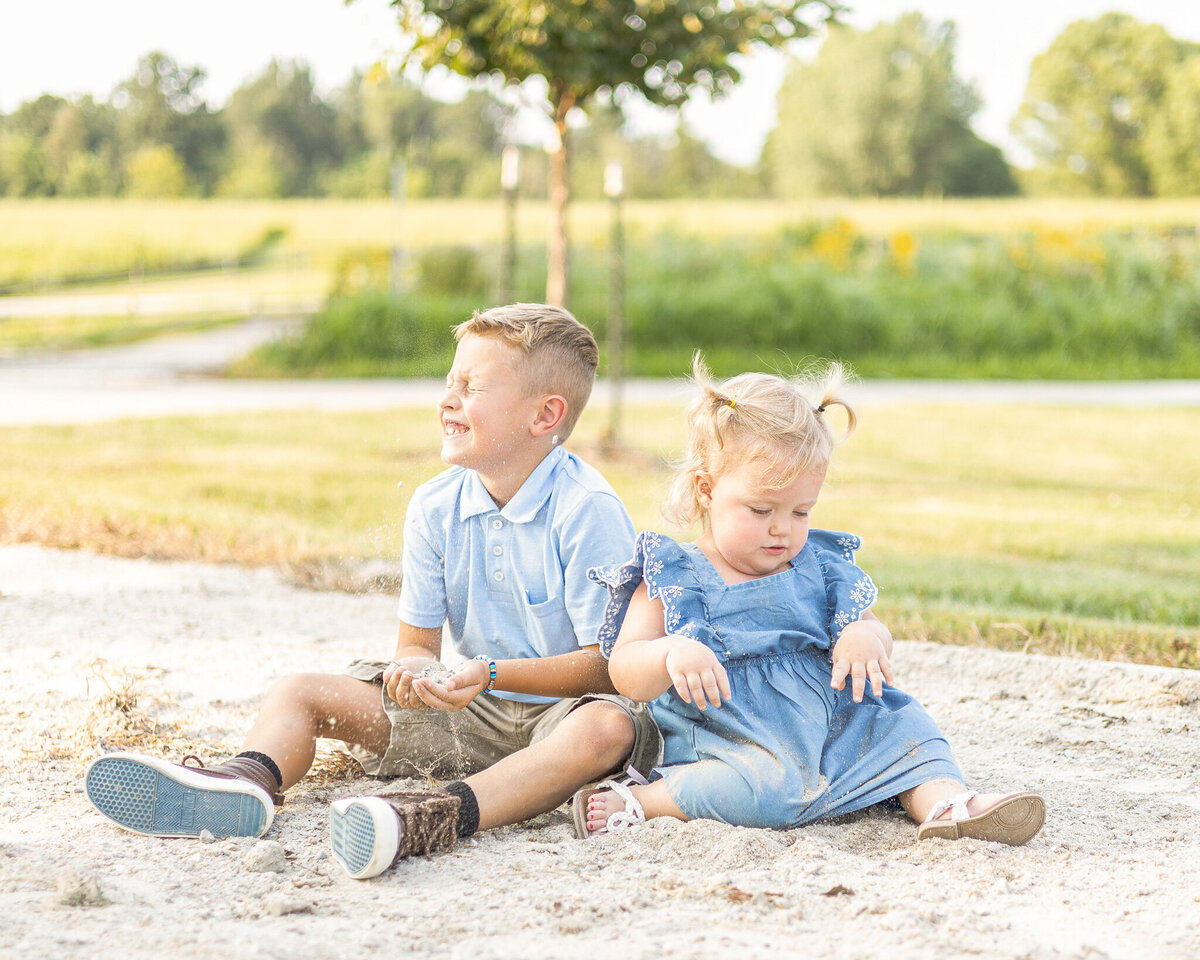 Young brother and sister sitting on the ground playing.  brother has short brown hair, wearing a partial button up short sleeve shirt, khaki shorts, brown shoes, with eyes closed looking away from the camera.  Sister has blonde hair in pigtails, wearing a blue dress, white sandals, and looking down at the ground