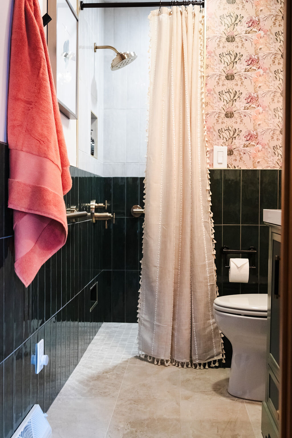 A bathroom featuring a shower area with dark green vertical tiles and a light gray mosaic tiled floor. A brass showerhead and faucet are visible. A partially drawn, extra-long, off-white shower curtain with decorative tassels runs along a curved rod, separating the shower from the rest of the bathroom. Beyond the curtain, a toilet and a section of the wall with dark green tiles on the lower half and floral patterned wallpaper in shades of pink and beige on the upper half are visible. A light switch is on the floral wallpapered section. A glimpse of a dark vanity with a white countertop is on the right. A coral-colored towel hangs on a brass fixture on the left.