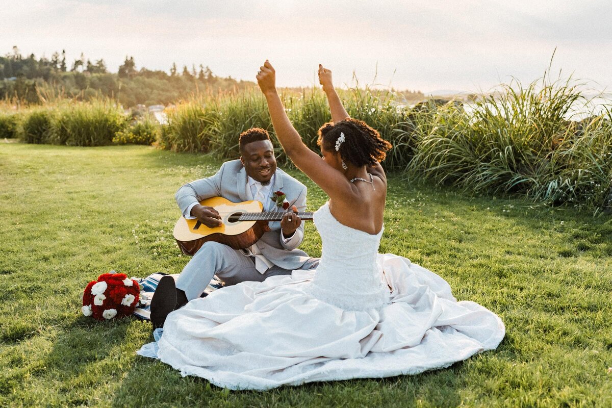 groom playing guitar and bride cheering him on while sitting in the grass next to the Washington Coast.