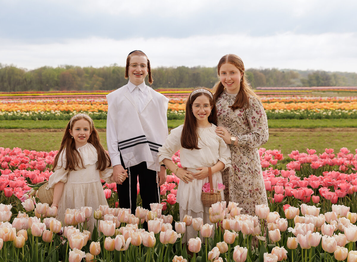 Brother and three sisters stand in a tulip field for a family photoshoot. All siblings are looking at the camera smiling. Captured by Chaya Bornstein, Brooklyn Family Photographer