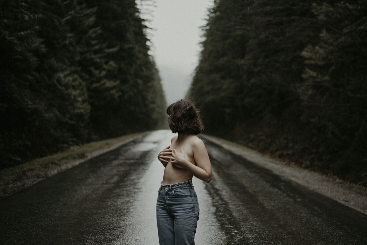 girl in the middle of a street through the forest, wearing jeans and cupping her topless breasts with her hands.