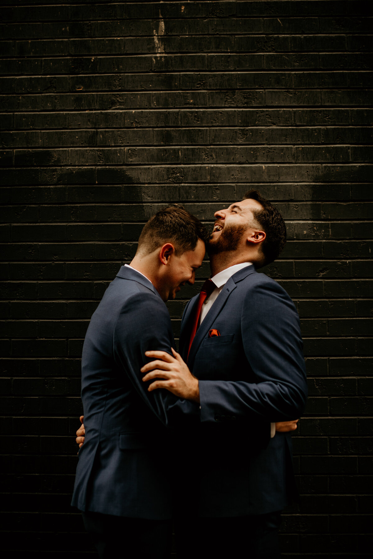 two grooms in blue suits hodling each other in front of a black brick wall in Albuquerque laughing together