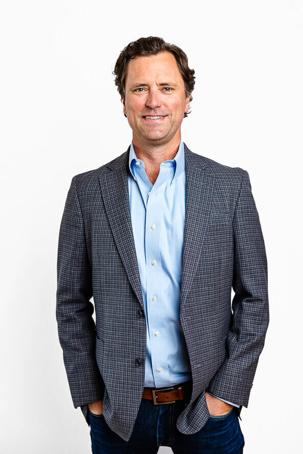 Man in blue shirt and jacket with hands in pockets poses in white studio.