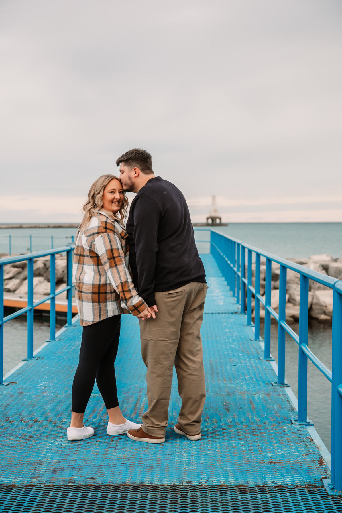 man kisses womans temple as she looks back at camera on a blue bridge over lake michigan in port washington