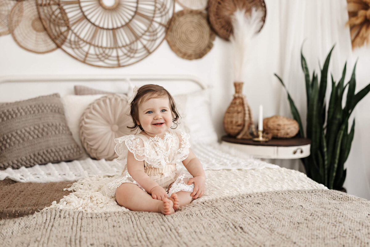 A smiling baby in a lacy, cream-colored outfit sits on a cozy, textured bed with neutral pillows. Woven decor and a green plant decorate the stylish, boho-inspired bedroom—perfect for milestone celebration portraits.